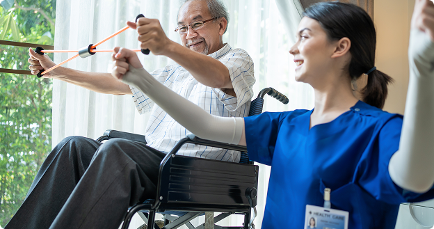 ChatGPT said: Caregiver in blue scrubs leads an exercise, demonstrating arm movements with a resistance band while an older man in a wheelchair follows along in a bright room.