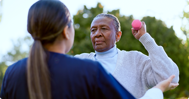 Older adult lifting a small pink dumbbell outdoors while a caregiver stands in front providing support and guidance.