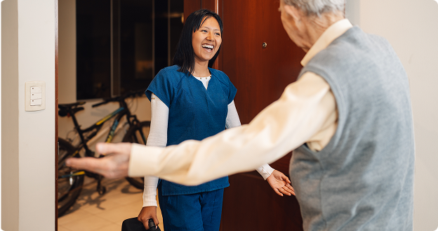 Smiling caregiver in blue scrubs arrives at a client’s home with a bag in hand, while an older man greets her warmly with open arms at the doorway.