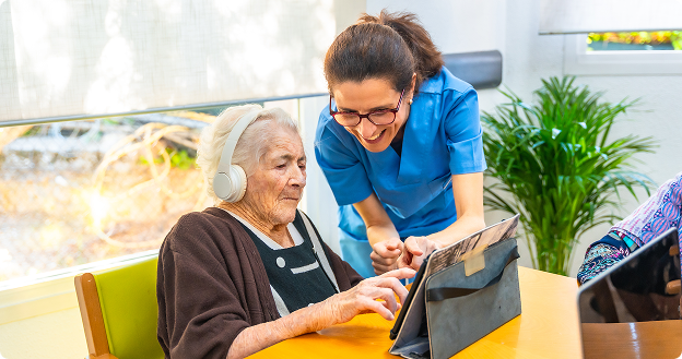 Older woman wearing headphones uses a tablet at a table while a smiling caregiver in blue scrubs leans in beside her, guiding her with the device.