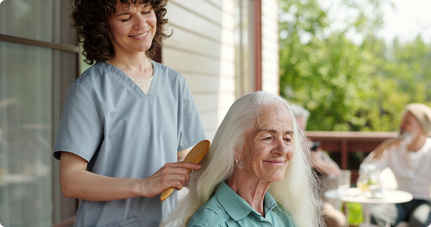 Smiling caregiver brushes the long white hair of an older woman sitting on a porch, with other residents relaxing at a table in the background.